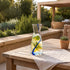 A clear glass carafe with a blue spiral stripe, filled with water and lemon slices, set on a wooden outdoor patio table.