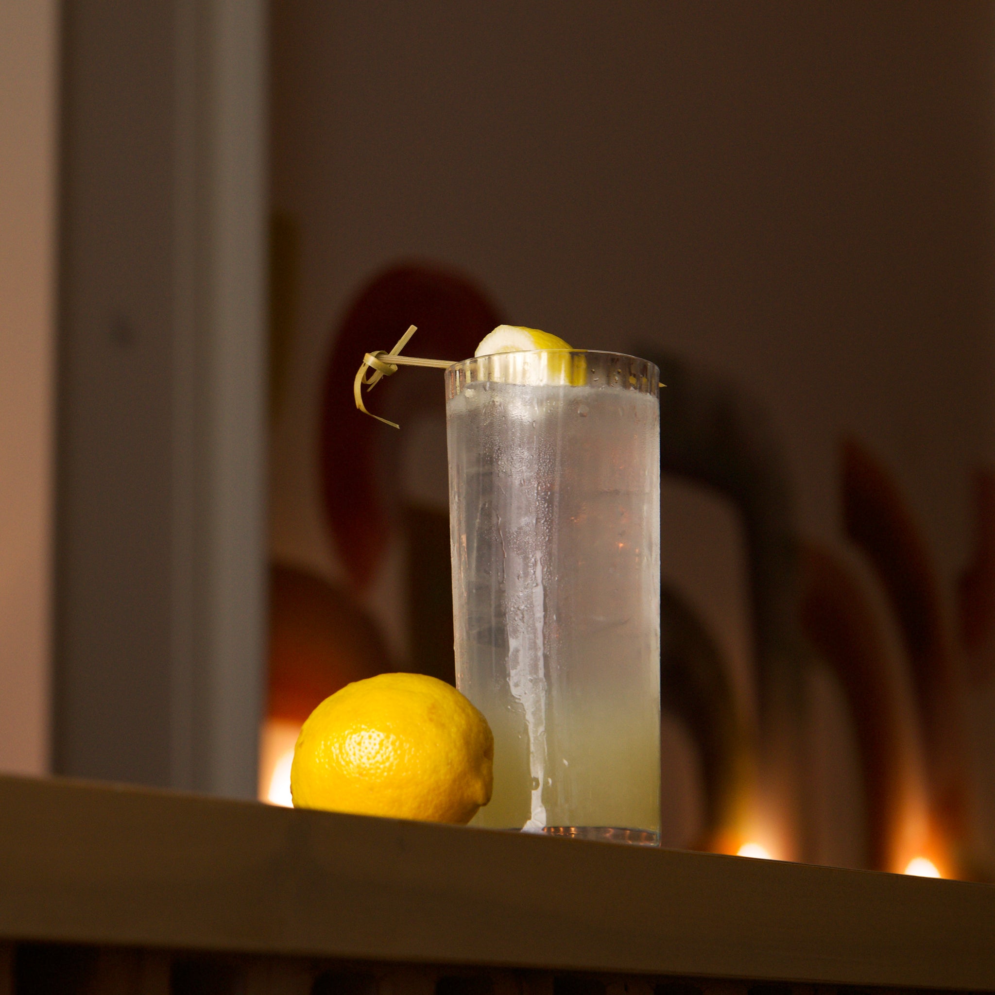 Clear Exclusiva Tom Collins glass filled with a light cocktail and garnished with a lemon peel on a wooden surface and a lemon lying next to the glass