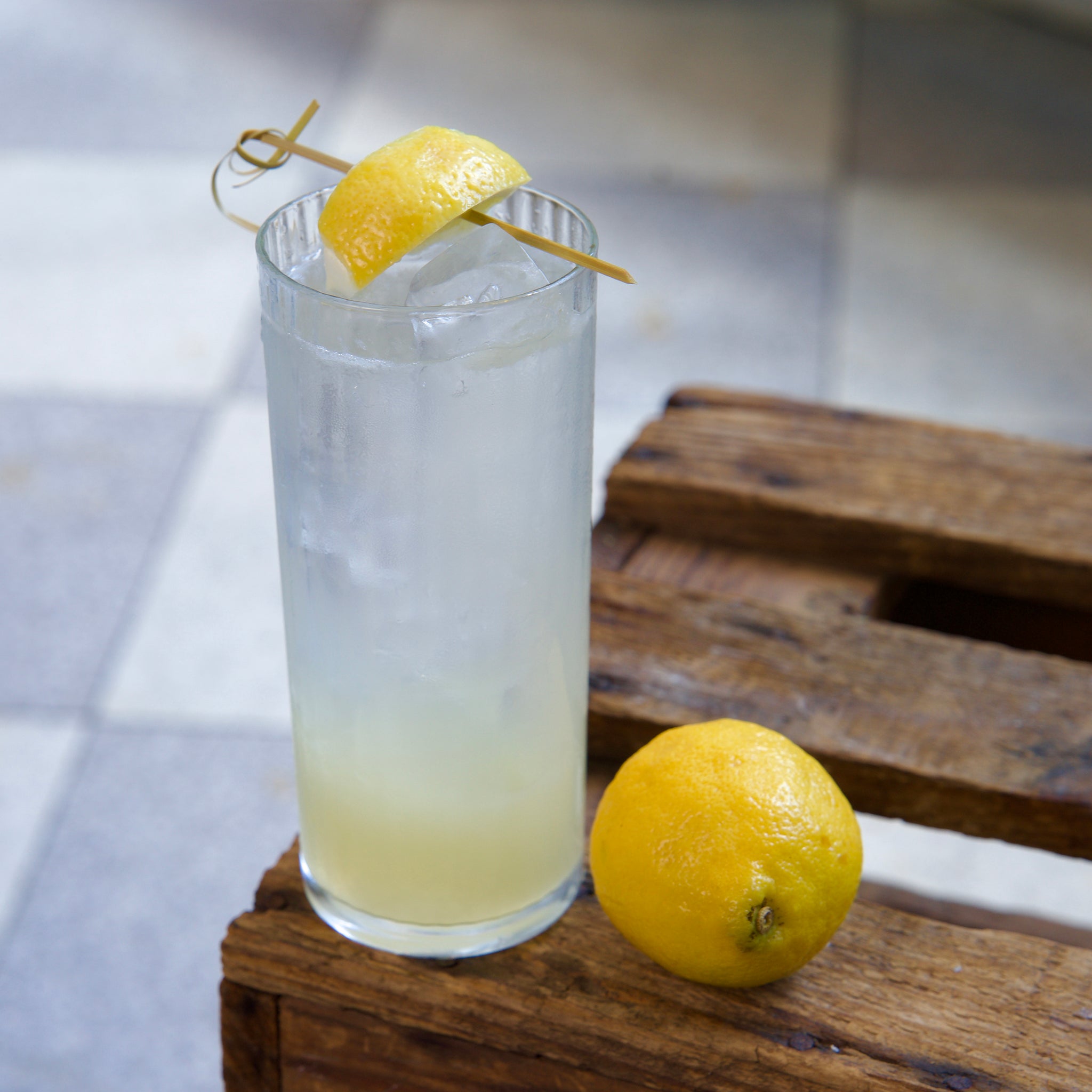 Exclusiva Tom Collins glass filled with a light cocktail with lemon garnish on a wooden crate with a lemon next to it