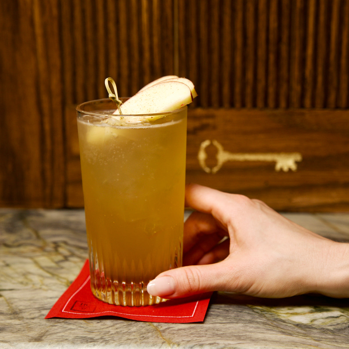 Graphics Baverage glass with a cocktail with apple garnish held by a hand on a wooden surface.