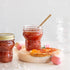 Two Quattro Stagioni special edition jars with red jam with a spoon on a wooden board with a bread slice.