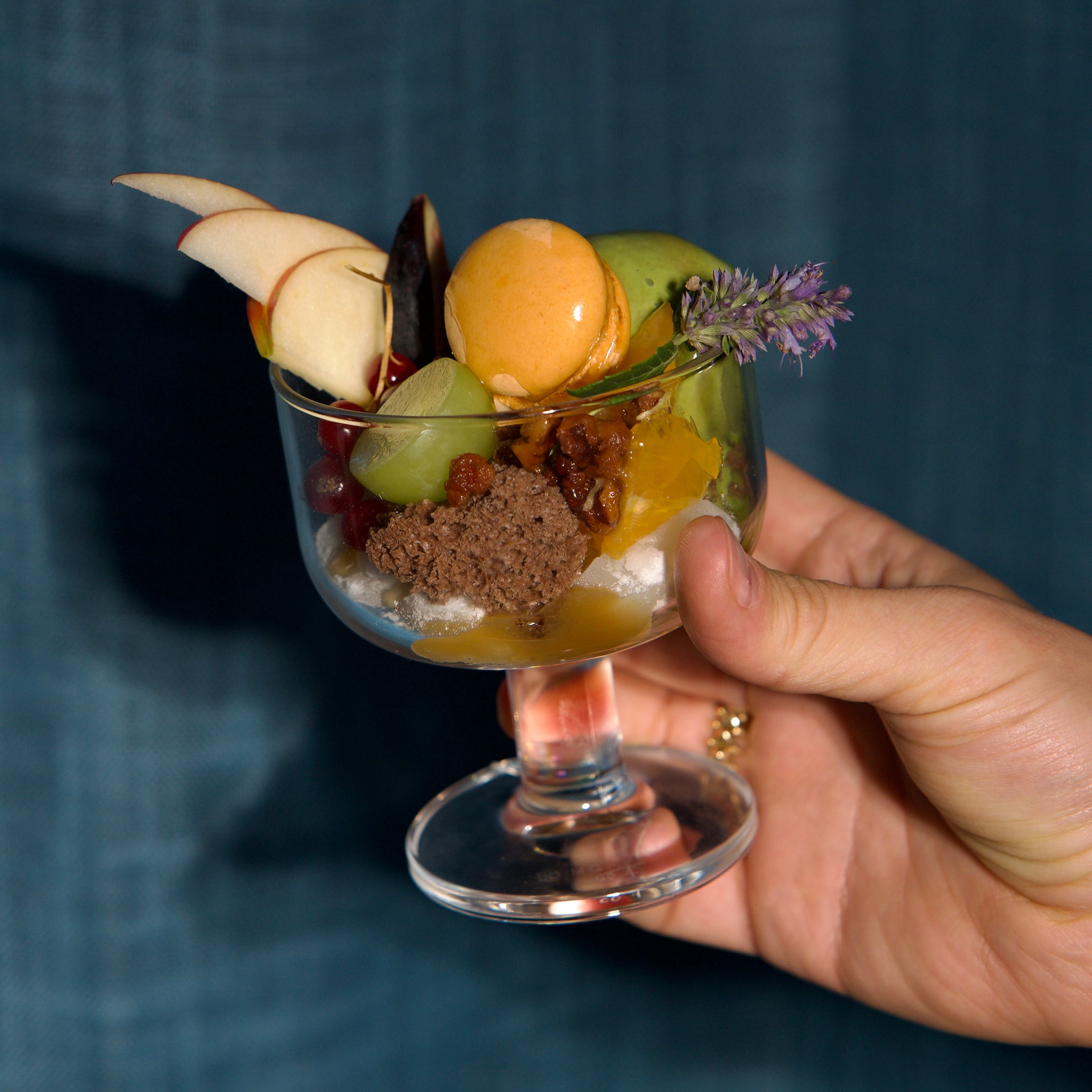 Hand holding a glass dessert with fruits and a flower against a dark background