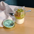 Small dessert jar with granola and fruit on a wooden table with a container of 'Quattro Stagioni' on a white cloth.