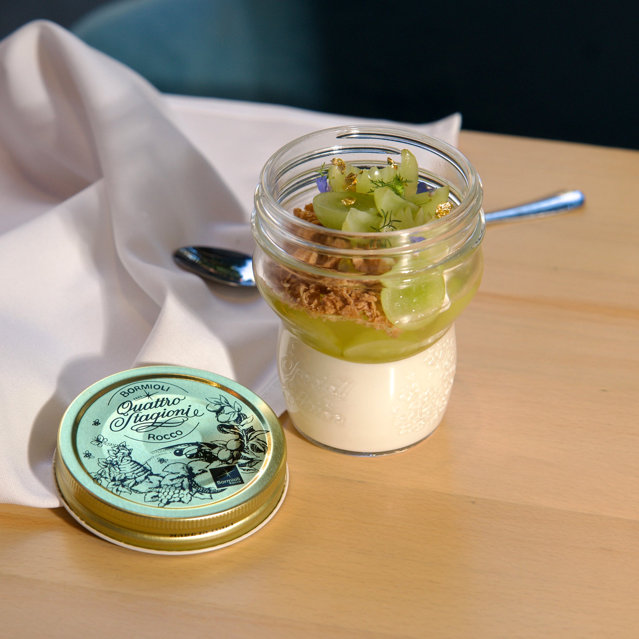 Small dessert jar with granola and fruit on a wooden table with a container of 'Quattro Stagioni' on a white cloth.